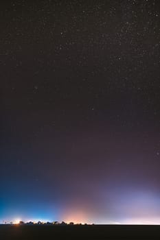 Night Starry Sky With Glowing Stars Above Countryside Town Landscape. Rural Field Meadow In Early Spring. Bright Glow Of Stars. Illumination Lights Of Small Town Or Village.