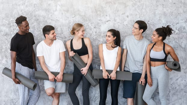 Healthy Lifestyle Concept. Group of sporty people with yoga mats resting after class near grey wall in fitness studio