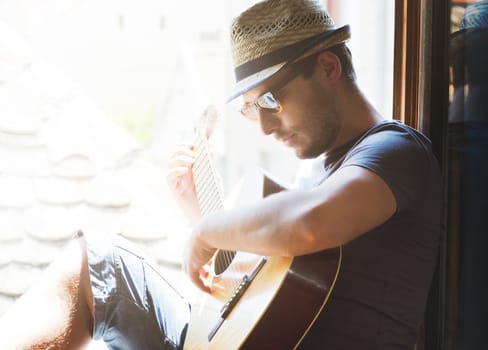 Portrait of young stylish guy with hat and sunglasses plays guitar at home. Creative man have leisure time with music at summer holiday. Hipster lifestyle concept.