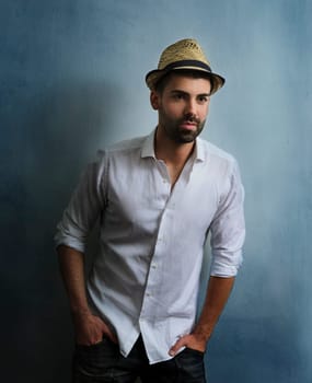 Young sexy hipster guy with beard posing in front of blue grunge wall. Portrait of stylish male model with retro hat and casual style clothing. Man's fashion and beauty.