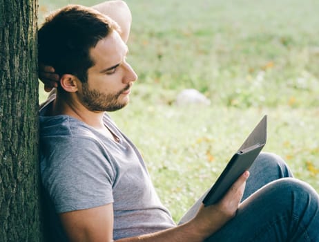 Young man sitting on the grass in nature and relaxing while reading from e-book reader.