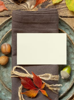 Autumn rustic table place with blank card between colorful leaves and berries, top view, mockup. Flat lay with horizontal card on wooden table, copy space. Thanksgiving or harvest concept