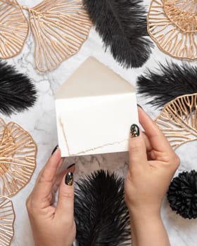 Hands with Blank card and envelope near black feathers and golden leave top view. Invitation or greeting card mockup. Elegant femminine flat lay with vertical paper card