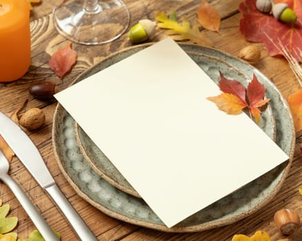 Autumn rustic table setting with blank card between colorful leaves and berries, close up. Vertical card mockup on wooden table, copy space. Thanksgiving, halloween or harvest concept