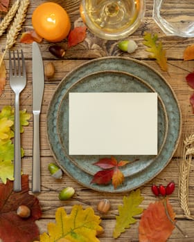 Autumn rustic table setting with blank card between colorful leaves and berries, top view. Horizontal card mockup on wooden table, copy space. Thanksgiving, halloween or harvest concept