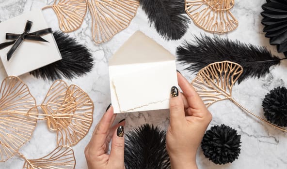 Hands with Blank card and envelope near black feathers and golden leave top view. Invitation or greeting card mockup. Elegant femminine flat lay with vertical paper card