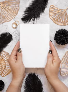 Hands with Blank card near black feathers, golden leaves and wedding rings top view. Invitation or greeting card mockup. Elegant femminine flat lay with vertical paper card
