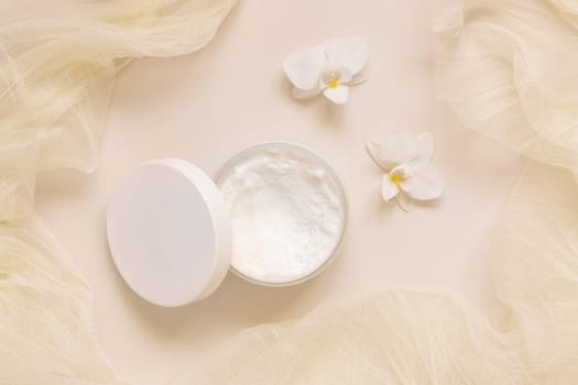 Opened Cream jar with a lid near white orchid flowers and tulle on light yellow top view. Cosmetic Mockup. Exotic natural cosmetics, romantic composition, everyday skincare routine..