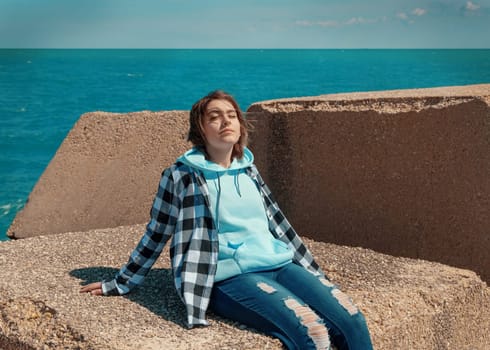 Relaxed Teenager girl in light blue hoodie and plaid shirt. Smiling Teen girl sitting outdoor and enjoying the sunny day. Adolescence concept