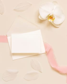 Wedding Card over envelope near white orchid flower and silk ribbons on light yellow top view, mockup. Tropical scene with horizontal blank card flat lay