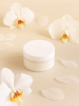 Cream jar with a white lid near white orchid flowers on light yellow close up. Cosmetic packaging Mockup. Exotic natural cosmetics, romantic composition, everyday skincare routine..