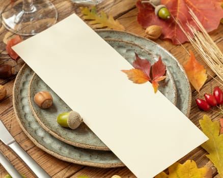 Autumn table setting with red, yellow and orange leaves, berries and blank card close up. Fall Mockup of vertical menu card on wooden table, copy space. Harvest, Halloween and Thanksgiving season