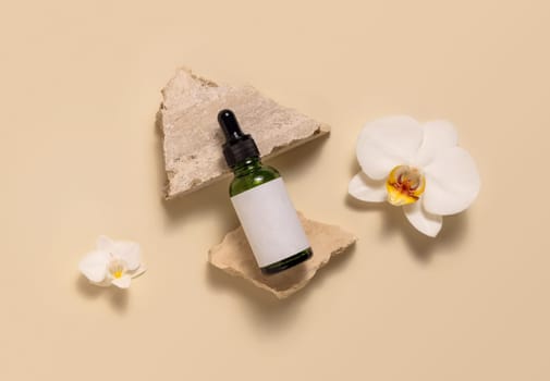 Green glass dropper bottle on stones near white orchid flowers on light yellow, top view, mockup. Skincare beauty product, serum or essential oil. Exotic natural cosmetics..