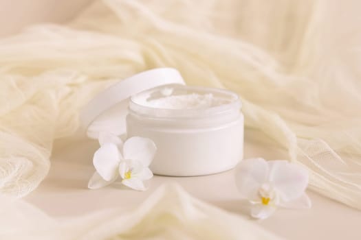 Opened Cream jar with a lid near white orchid flowers and tulle on light yellow top view. Cosmetic Mockup. Exotic natural cosmetics, romantic composition, everyday skincare routine..