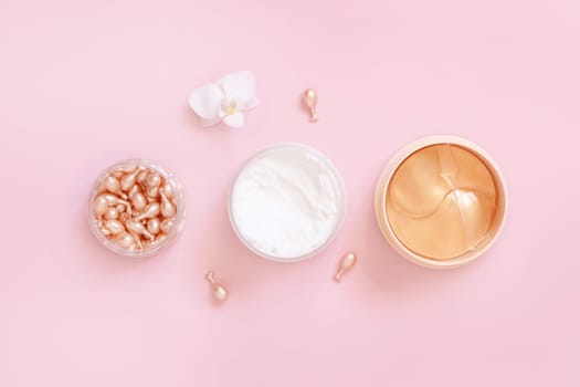 Face serum capsules, cream and eye patches on light pink near orchid flower top view. Skincare cosmetics. Facial Beauty treatments. Pastel minimal composition..