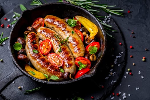 Grilled sausages and vegetables with herbs in iron pan on black background