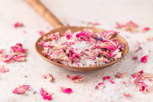 See salt wih with dried flower petals in wooden scoop on pink marble table