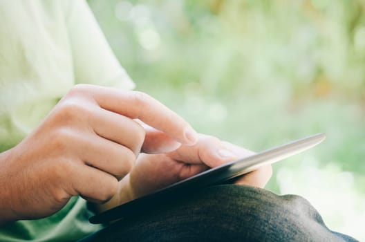 Closeup of hand holding computer tablet with green natural bokeh background. Young man using tablet device for e-leaning, education, shopping, leisure or business while is in the park.