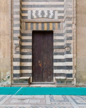 Wooden aged door surrounded by striped black and white marble decorations and stone wall at the courtyard of Sultan Hassan Mosque, Cairo, Egypt