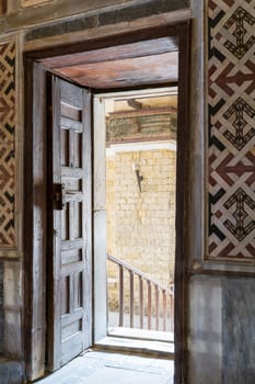 Angled view of a wooden aged ornate opened door leading to a passage with bright light, color decorated marble wall, and marble floor at historic public mosque of Sultan Qalawun, Cairo, Egypt
