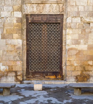 Wooden grunge window with decorated iron grid over stone bricks wall and two marble garden benches, Moez Street, Cairo, Egypt