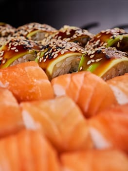 Close up of sushi rolls with slices of avocado, sesame and sauce with California rolls in a blurred foreground. Selective focus.