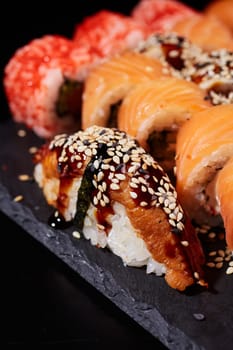 Close up of traditional nigiri sushi with tuna, sauce and sesame seeds lying on the edge of a black stone plate on a background of other types of rolls. Selective focus. Blurred background.