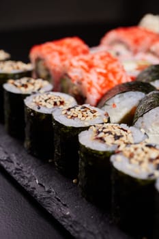 Close up of sushi with sesame seeds wrapped in nori leaves lying on a black board on the background of other types of sushi. Blurred background. Selective focus.