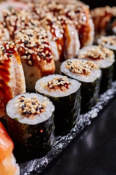 Close up of maki sushi with tuna, sauce and sesame seeds lying on the edge of a black stone plate. Selective focus. Blurred background.
