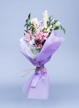 Bouquet of fresh natural flowers wrapped in floral tracing paper stands in a vase on a blue background. Bouquet in pink style composed of irises, other different flowers and twigs of cotton.