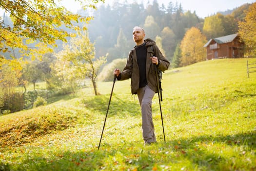 Portrait of a young male tourist with a backpack on his shoulders and trekking sticks in his hands. Man travels through the mountains, stands on a mountainous countryside and looks away.