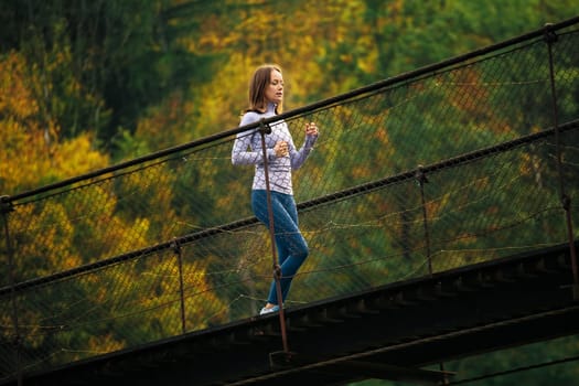 View from below of a young sports woman performing a morning jog on a suspension bridge against the backdrop of a mountain forest. Concept of active and healthy lifestyle. Blurred background.