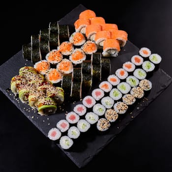 Assortment of different kinds of sushi rolls placed on black stone board. Sushi set with salmon, cucumbers, cream cheese, avocado and sesame seeds. Sushi image for menu.