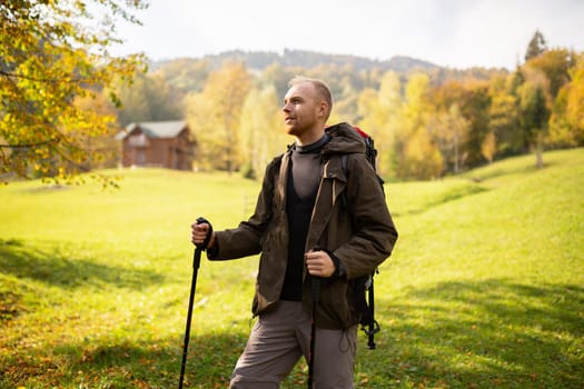 Young male traveler with a backpack and trekking sticks in his hands decided to stop to relax before a walk in the mountains. Man looks away and enjoys the natural scenery in the light of the sun.