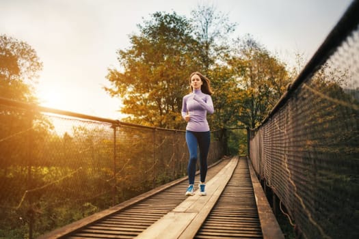 Portrait of a young woman in sportswear while jogging on a suspension bridge. Fitness woman working out in nature on a cool autumn day. Outdoor cardio exercise concept.