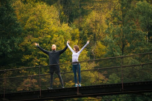 Healthy lifestyle. Young man and woman standing with arms raised on a suspension bridge on a background of forest. Sports couple warms up before jogging in the mountains.