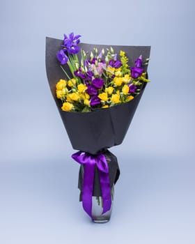Stylish bouquet of yellow and purple flowers wrapped in black paper, stands in a vase on a blue background. Festively packed bright colored bouquet of yellow and purple eustoma and irises.