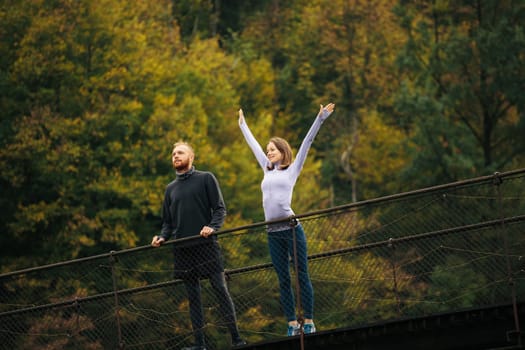 Mountain forest landscape. Young happy man and woman with joyful facial expression standing on a suspension bridge. Athletes in sports uniform are going to train outdoors. Concept of unity with nature