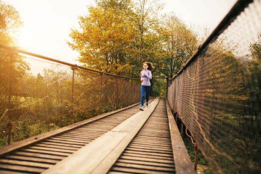 Young woman spends time with benefit. Female runner begins to jog over a wooden suspension bridge in the middle of a mountain forest. Fitness woman working out in nature on a cool autumn day.