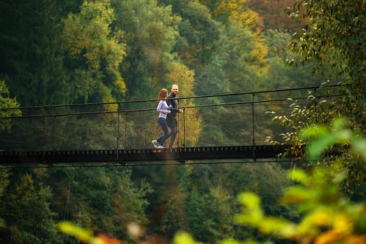 Mountain landscape. Side view of a young man and woman jogging on a mountain suspension bridge. Athletes in sportswear train outdoors. Young family spends time with benefit.