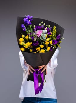 Bouquet of yellow roses and purple eustoma in a black paper wrapper in the hands of a woman standing on a gray background. Unknown woman holds out flowers in front of her, covering her face with them.