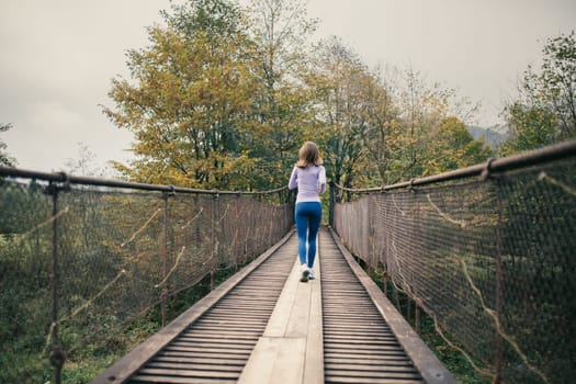 Rear view of a young athletic woman running across a suspension bridge during cardio exercises. Fitness woman working out in nature on a cool autumn day. Concept of active and healthy lifestyle.