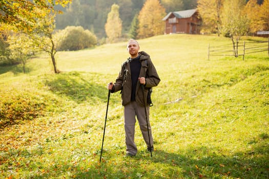Walking on top of the mountains. Male hiker with hiking equipment stands in the middle of a mountain village on a hill and looks at the camera. Happy man exploring the sunny mountains. Wanderlust.