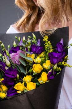 Close up of a stylish bouquet wrapped in black paper in the hands of an unrecognizable blonde woman. Bouquet is composed of purple eustoma and yellow roses. Birthday or Mother's Day concept.
