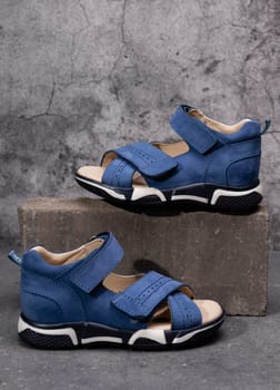Children's fashion. Pair of stylish blue children's sports sandals for boys on gray concrete background. Close up of one shoe standing higher on brick next to another which is lower.