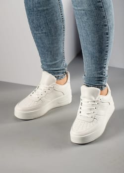 Casual comfortable white sneakers on female model in studio. Sports shoes with laces on feet of unknown young woman who is wearing jeans and standing on gray floor.