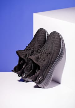 Black sneakers on blue background with white podium in form of geometric cube. Side view of unisex style sneakers standing leaning on podium. Spring and summer fashion concept.