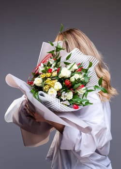 Woman in a delicate white shirt holds a bouquet of flowers with roses, tulips and greenery. Unknown blonde woman stands sideways on a gray background and holds flowers that cover her face.