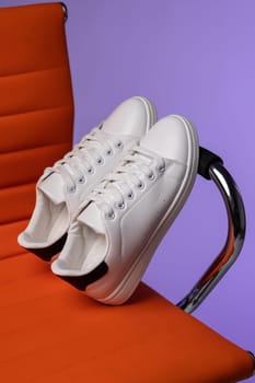 Creative promotion. Advertising photo of white women's or teen sneakers on chair in studio. Close up of white sneakers with black inserts standing leaning on handle of red chair on purple background.