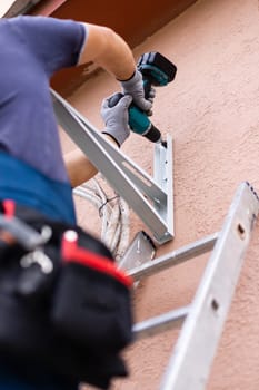 Installation of external block of conditioner by means of drill for fastening of holders of block. Worker standing on ladder attaches air conditioning holder to outside wall of house. Selective focus.